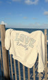 An off-white crewneck sweatshirt with 'LOVE Loudly' printed on the front, draped over a wooden beach fence with the ocean and blue sky in the background.