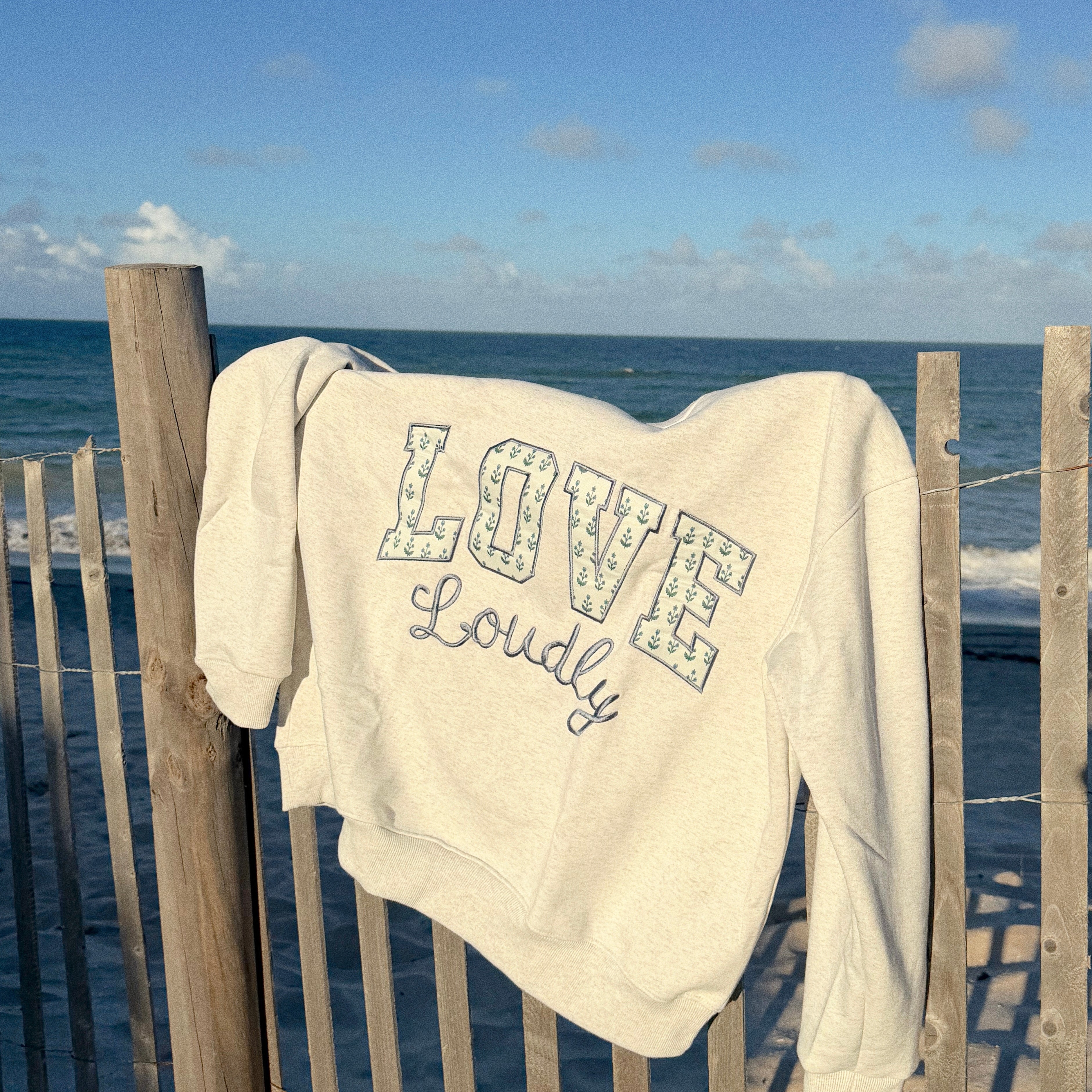 An off-white crewneck sweatshirt with 'LOVE Loudly' printed on the front, draped over a wooden beach fence with the ocean and blue sky in the background.