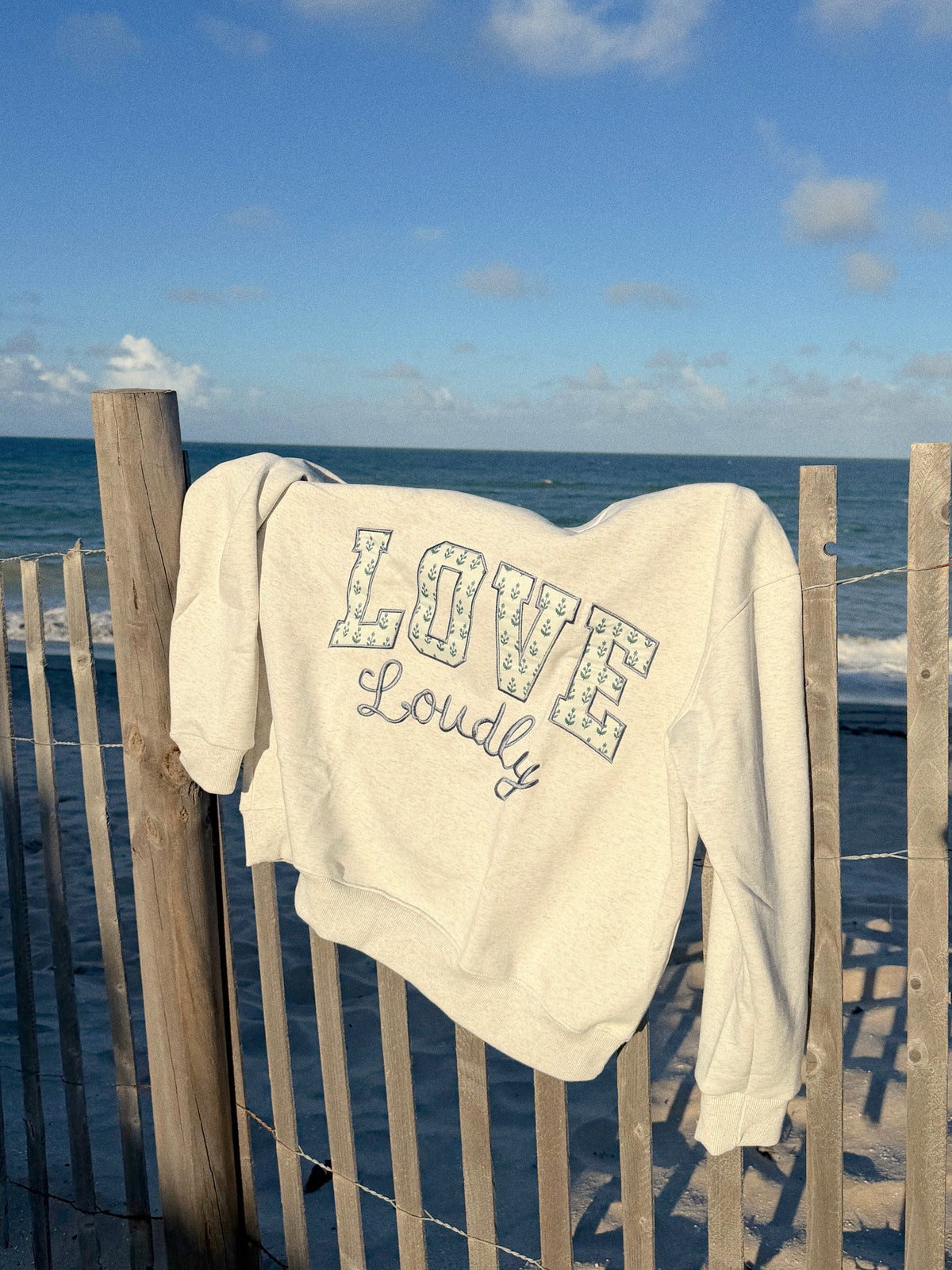 An off-white crewneck sweatshirt with 'LOVE Loudly' printed on the front, draped over a wooden beach fence with the ocean and blue sky in the background.