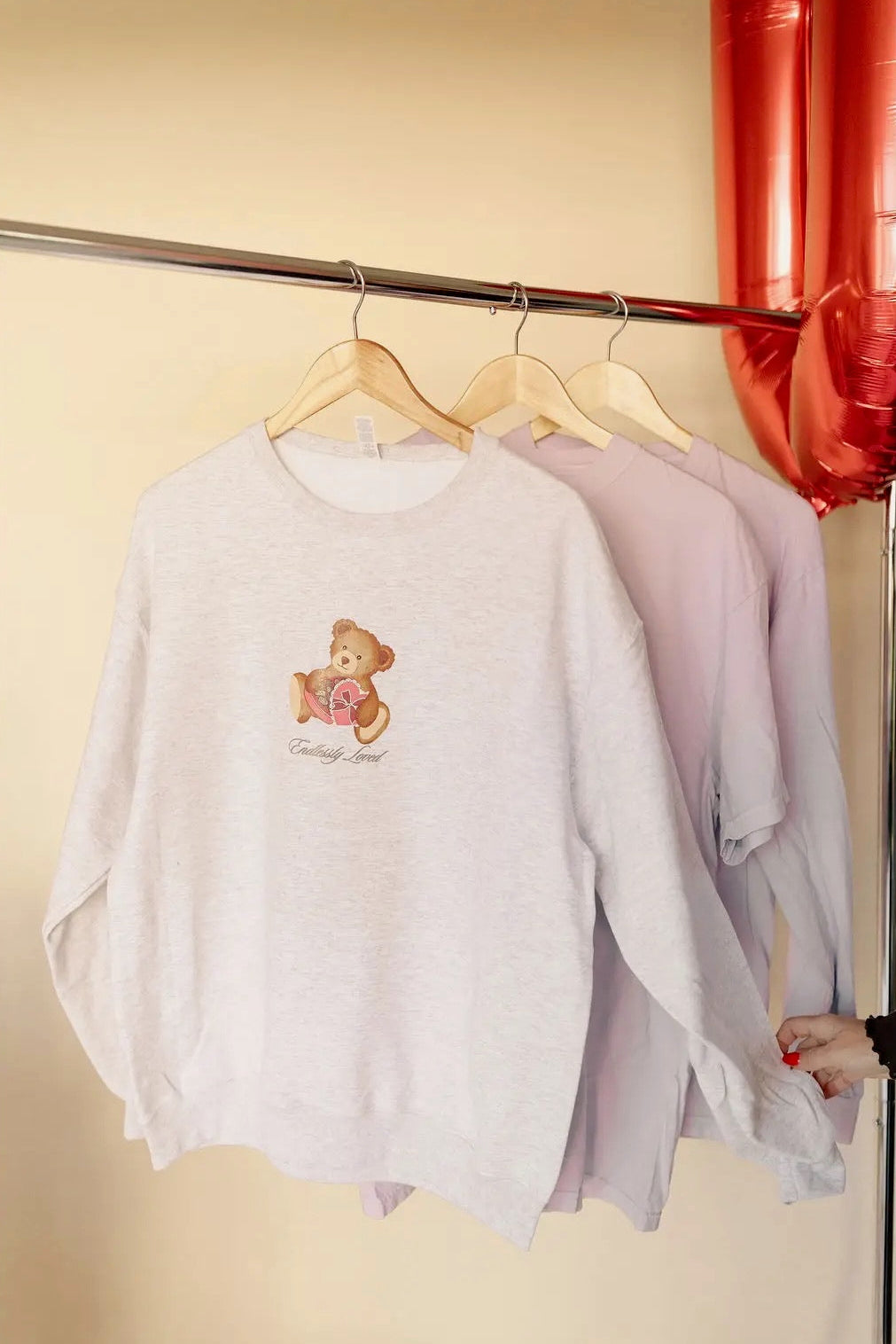 Light gray sweatshirt with teddy bear design that says "endlessly loved" on a hanger against a yellow wall. Valentine's Day themed
